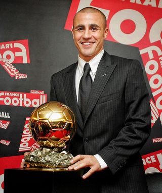 Fabio Cannavaro vestindo terno e gravata pretos, posando ao lado do troféu bola de ouro da Fifa